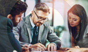 An accounting executive signing documents for two executive assistants seated at a desk.